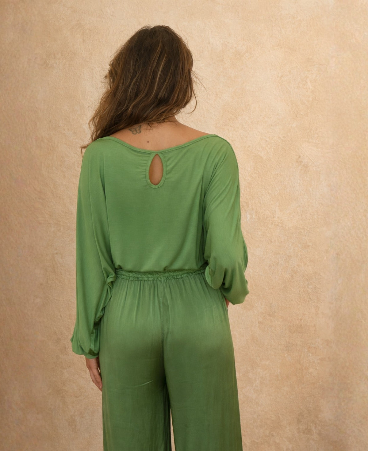 Back view of green satin keyhole top featuring teardrop keyhole opening and soft boat neckline, shown with matching satin pants against Mediterranean limewash wall.