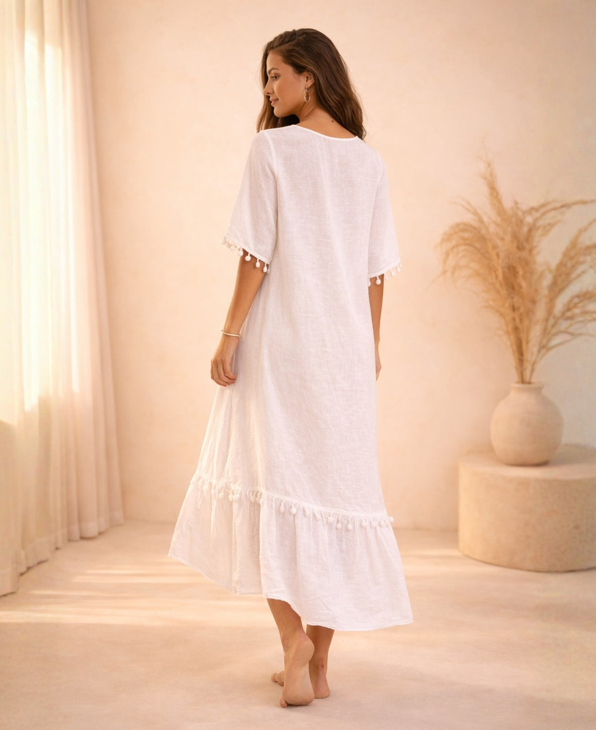 Back angled view of the Paradiso Kenya Linen Dress in white, showing the loose silhouette, higher back neckline, tassel-trim sleeves, and tiered hem in soft studio lighting.