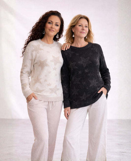 Two women wearing beige and black Apollo star sweaters standing together in a minimalist studio setting