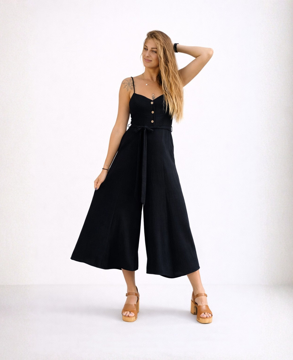 Front view of the Bella Byron Linen Jumpsuit in black, showing the structured bodice, decorative button placket, adjustable straps, waist tie belt and cropped palazzo leg in a minimalist studio.
