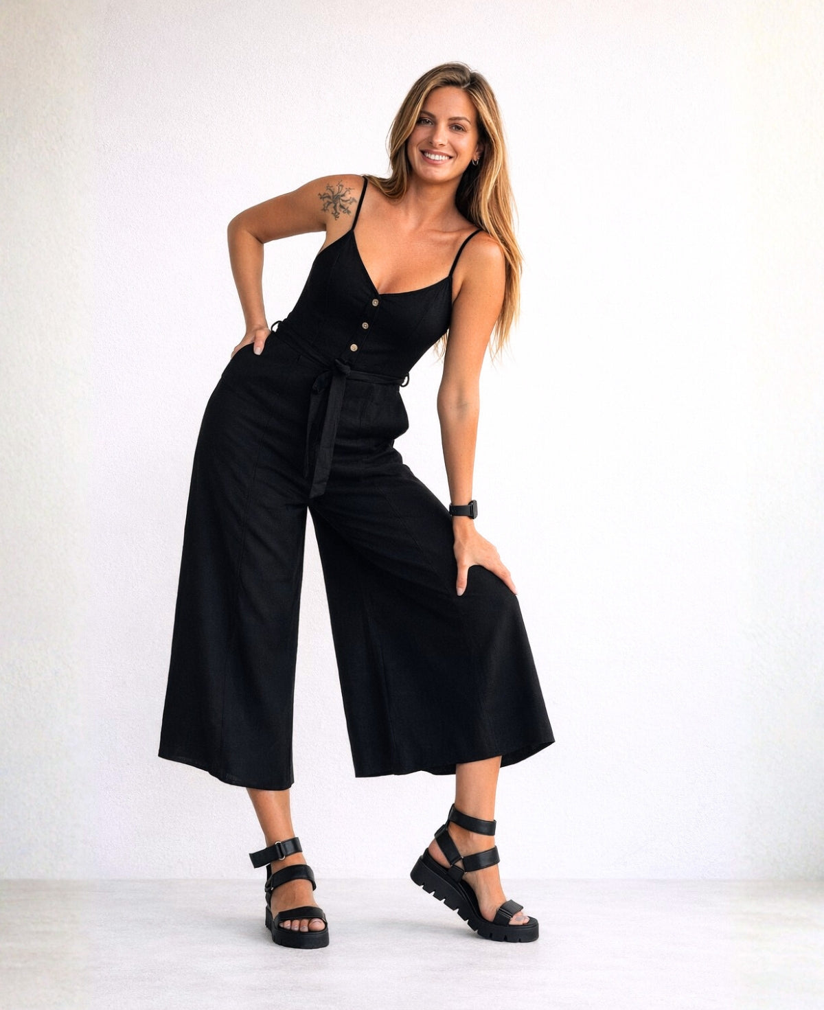 Model wearing the Bella Byron Linen Jumpsuit in black in a studio setting, posed with hand on knee to show fabric texture, front seams, tailored waist and wide cropped palazzo pants.