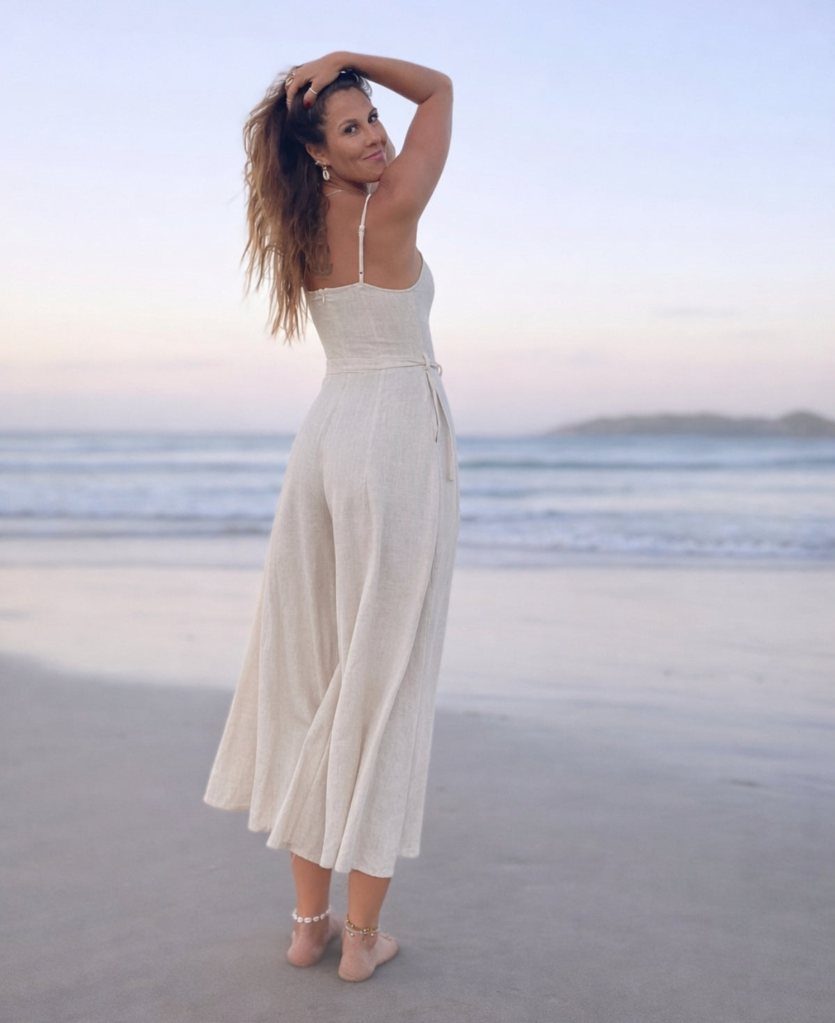Back view of the Bella Byron Linen Jumpsuit in ivory, showing the fitted bodice with back zip, adjustable straps, waist tie belt and wide cropped palazzo pants on a beach setting.