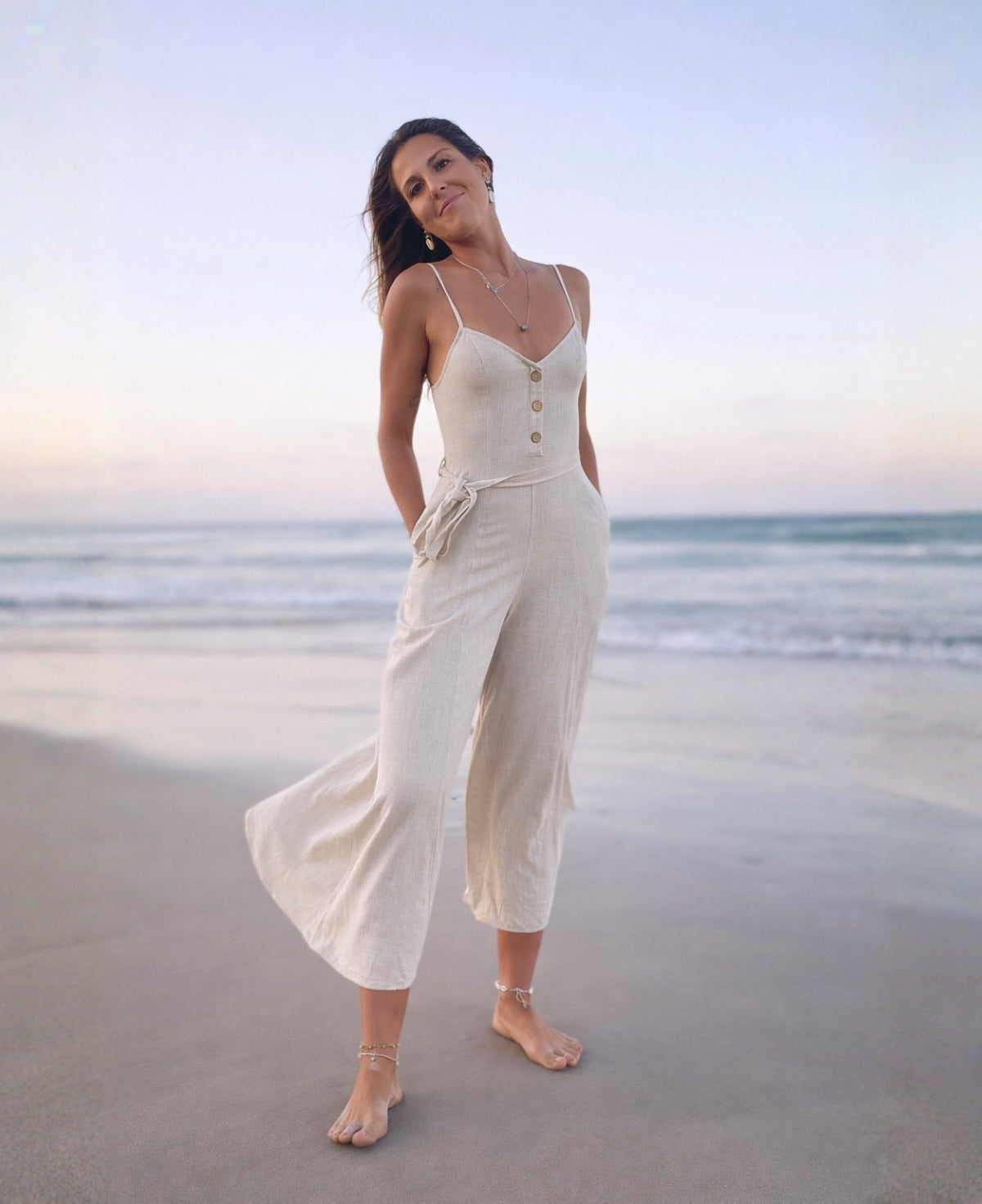 Model wearing the Bella Byron Linen Jumpsuit in ivory, shown from the front on the beach, featuring a structured bodice, button placket, adjustable straps, belt tie waist and cropped palazzo legs.