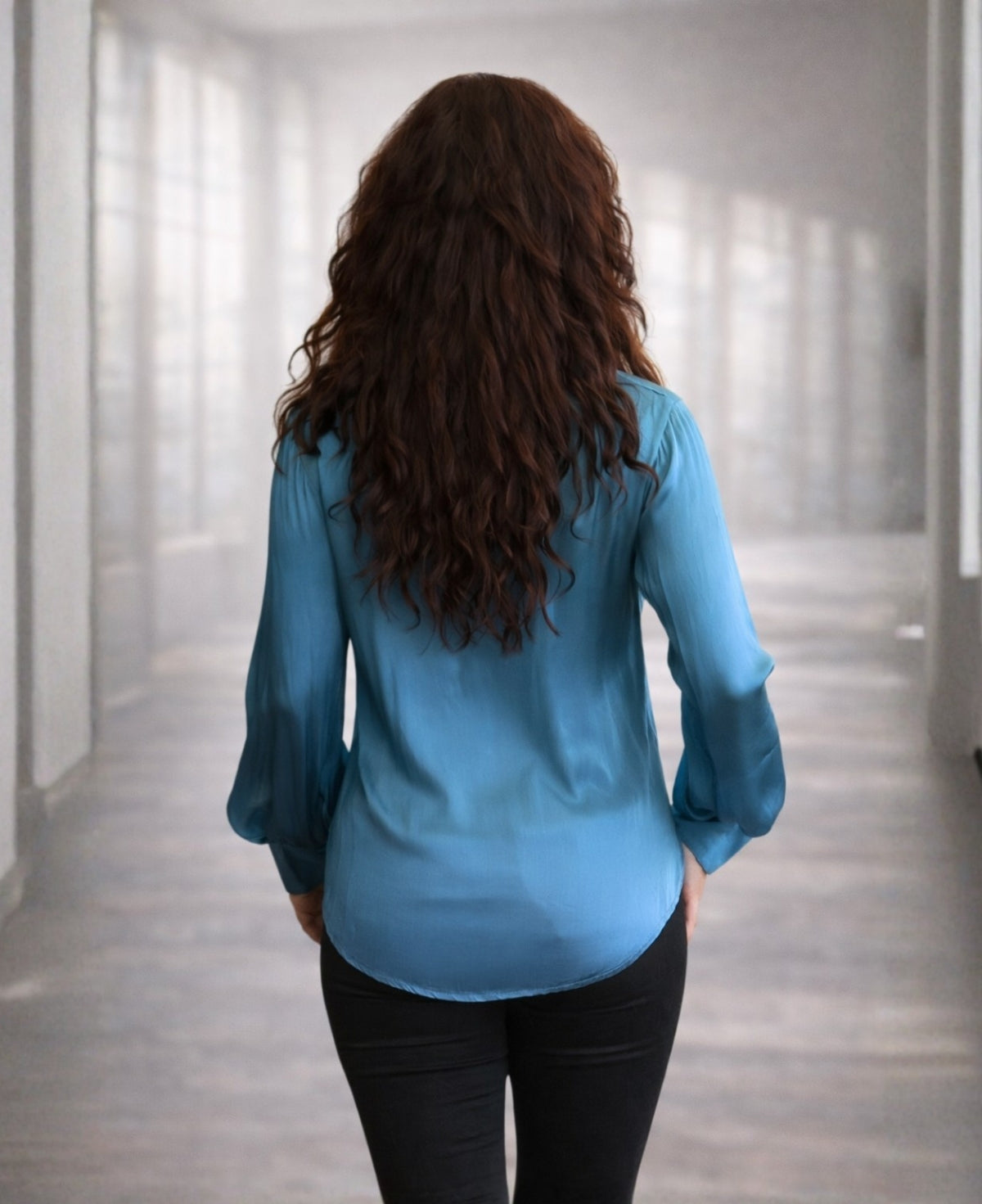 Back view of a blue teal satin button-up shirt showing the relaxed drape and smooth fabric, styled with black pants.