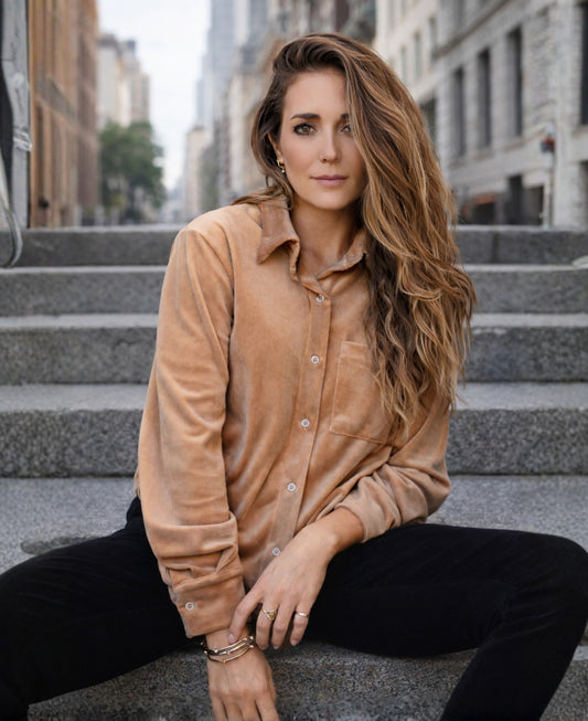 Woman wearing a camel velour button-up shirt sitting on outdoor city stairs, styled casually in an urban setting with a relaxed editorial feel.