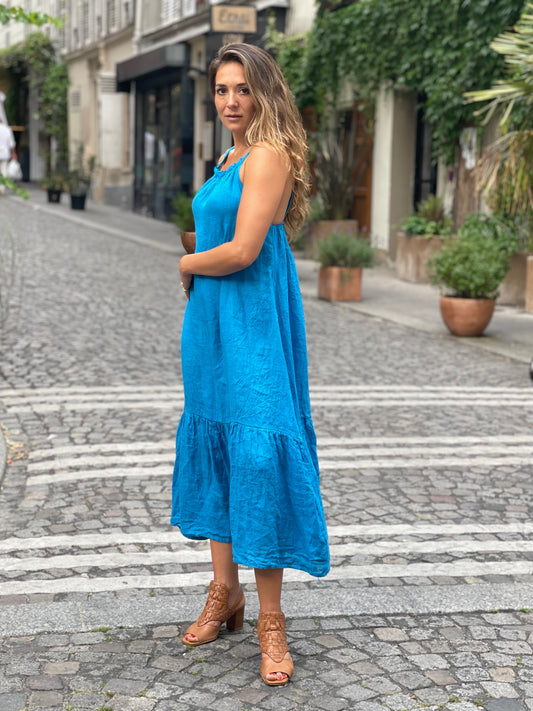 A woman wearing a turquoise linen midi dress with a ruffle hem and low open back, standing on a cobblestone street with greenery and buildings in the background.