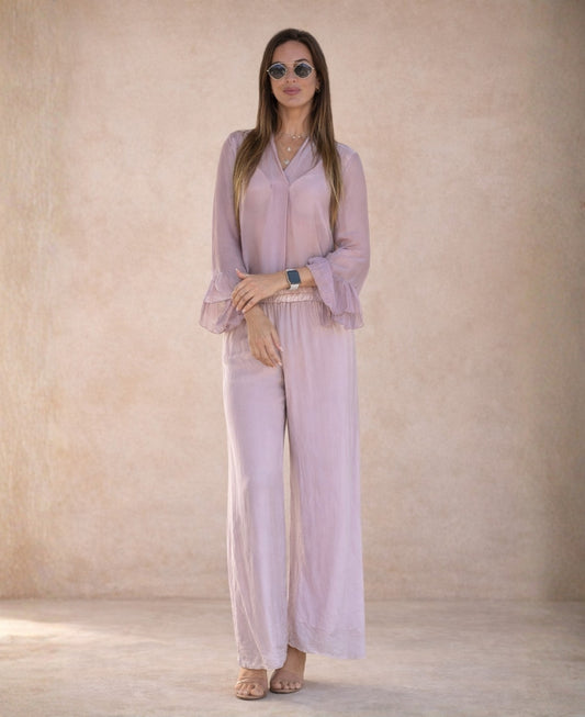 Model wearing blush silk blend pants with elasticated waist and wide leg fit, styled with a sheer blouse against a soft Mediterranean studio background.