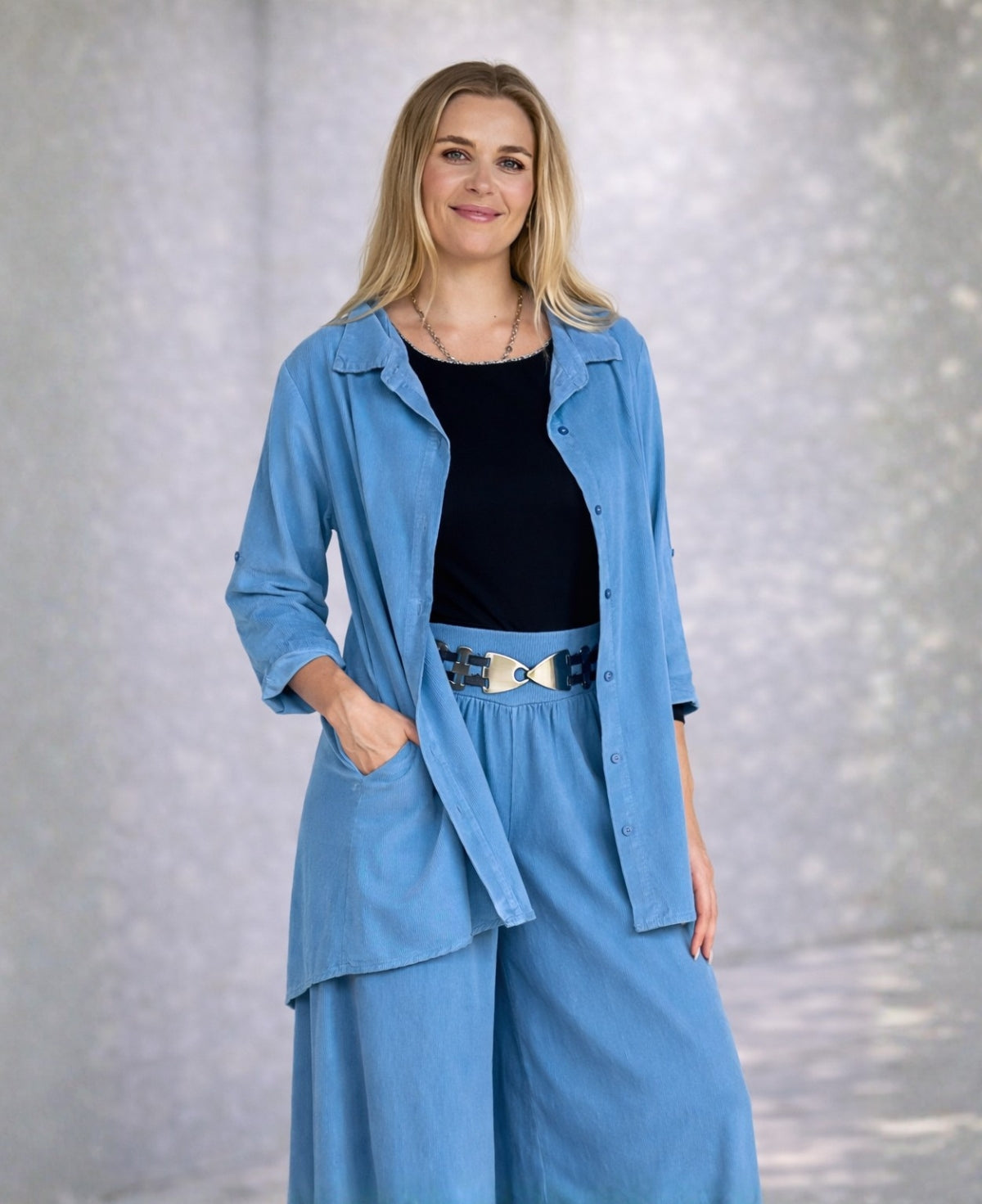 Model wearing the Dawn Corduroy Shirt in Blue Haven with fine ribbed corduroy texture, styled with matching pants and a black top, photographed against a soft neutral studio background.