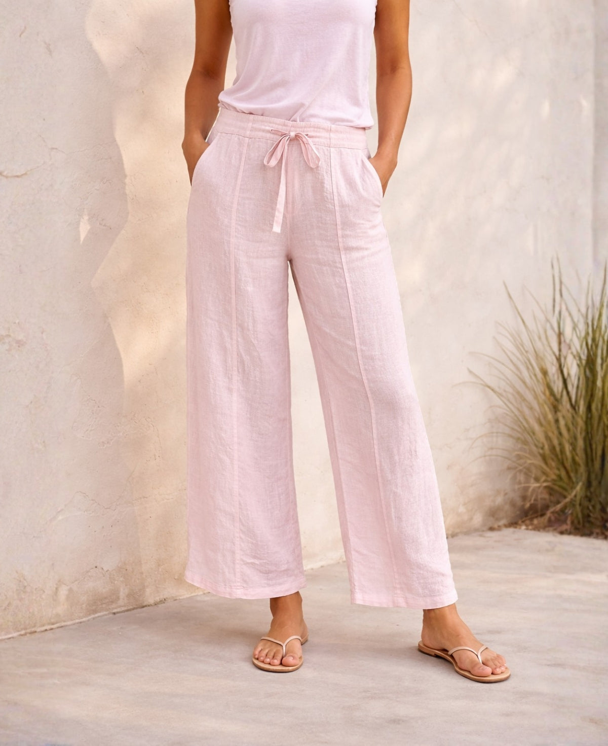Front view of model wearing Florence Linen Palazzo Pants in Blush, featuring wide-leg palazzo cut, visible front seams, elastic stretch waistband and matching linen tie, styled with nude sandals against a sun-washed Mediterranean wall.