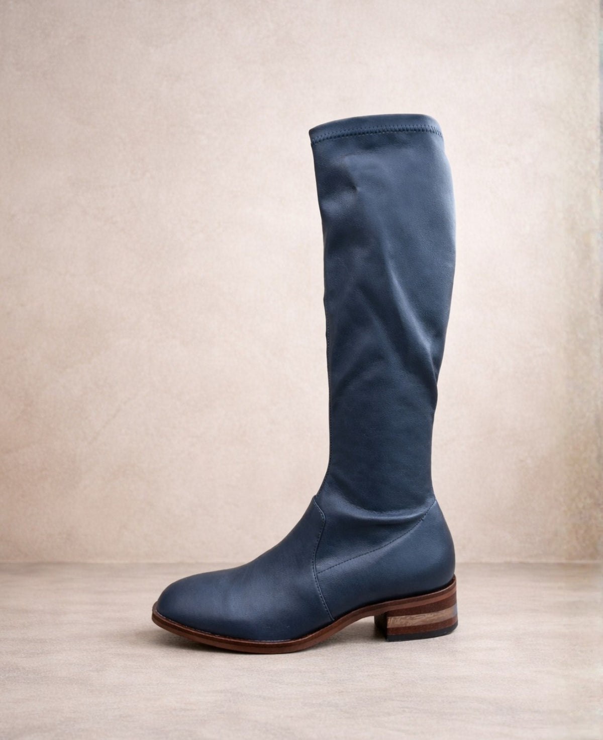 Side view of navy knee-high stretch boots with square toe and stacked heel on a warm neutral background.