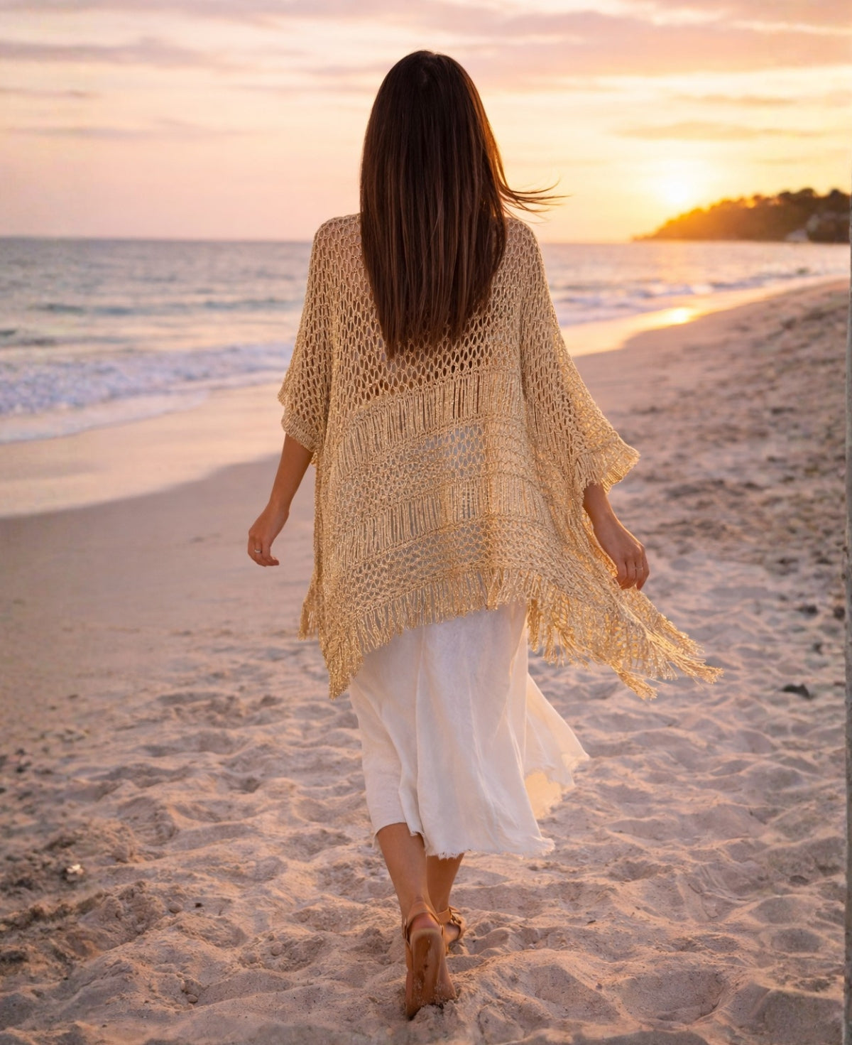 Back view of gold lurex crochet poncho with tassel trim flowing in the wind at sunset on the beach
