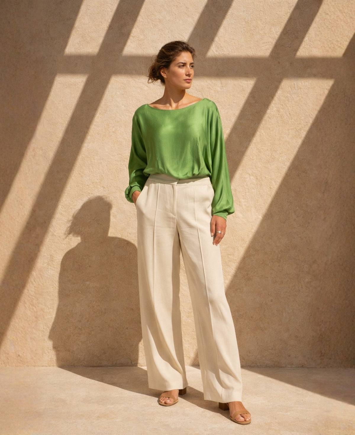 Editorial full body image of green satin keyhole top styled with cream wide-leg trousers and tan sandals, photographed against sunlit Mediterranean stucco wall with bold architectural shadows.