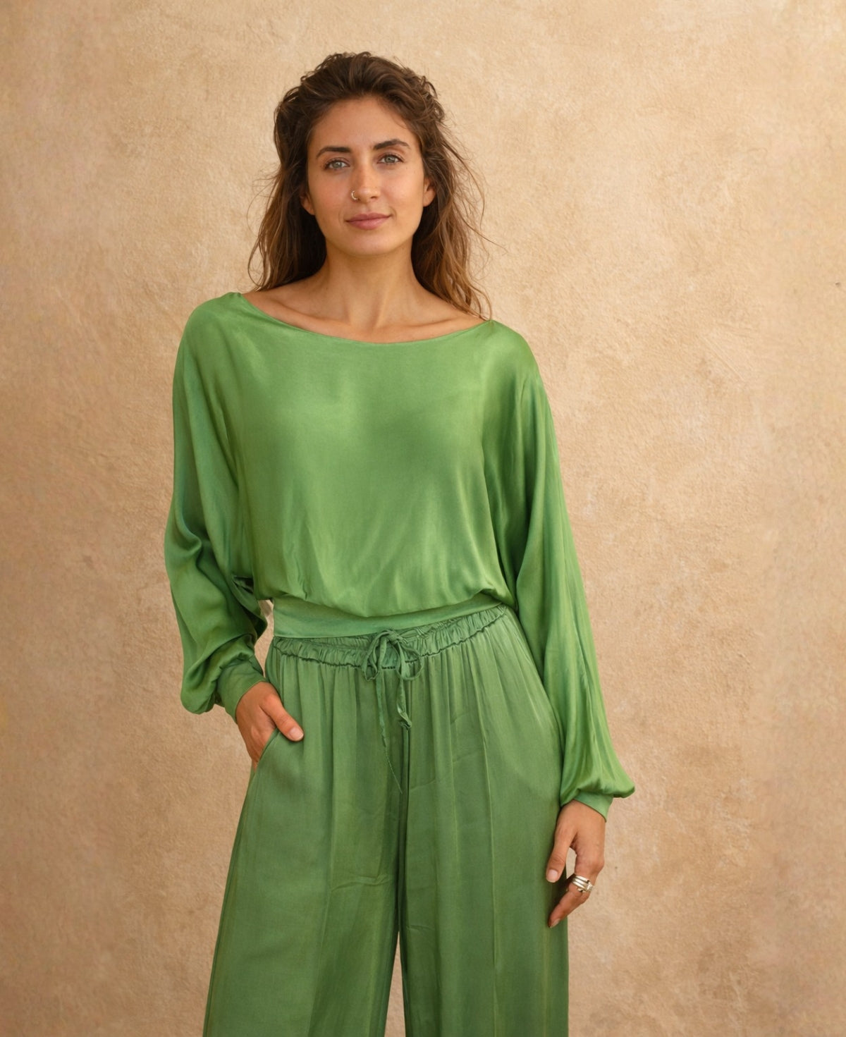 Front 3/4 view of green satin keyhole top with long sleeves and elastic waist detail, styled with matching satin wide-leg pants against warm sand textured background.