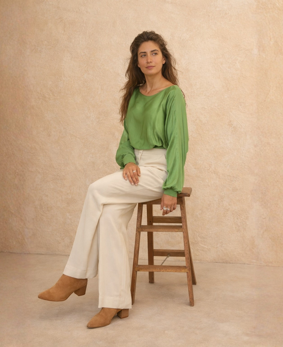Full body seated shot of green satin keyhole top styled with cream tailored wide-leg trousers and tan boots, photographed against warm sand studio wall.
