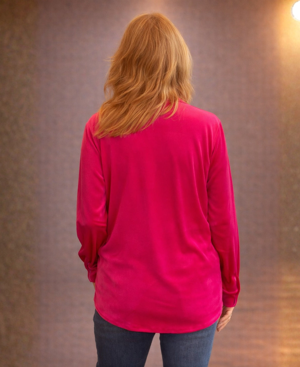 Back view of a hot pink velour shirt showing relaxed fit and drape, styled with jeans in a warm minimalist studio background.