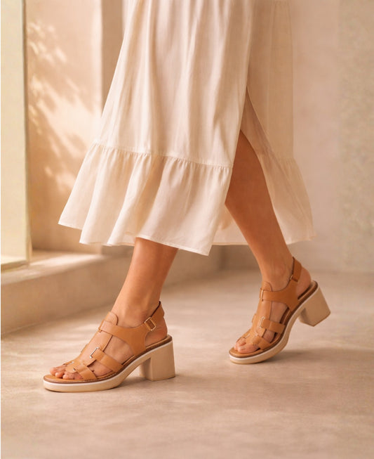 Woman wearing Jennis tan leather block heel sandals styled with a soft dress in a warm neutral studio setting