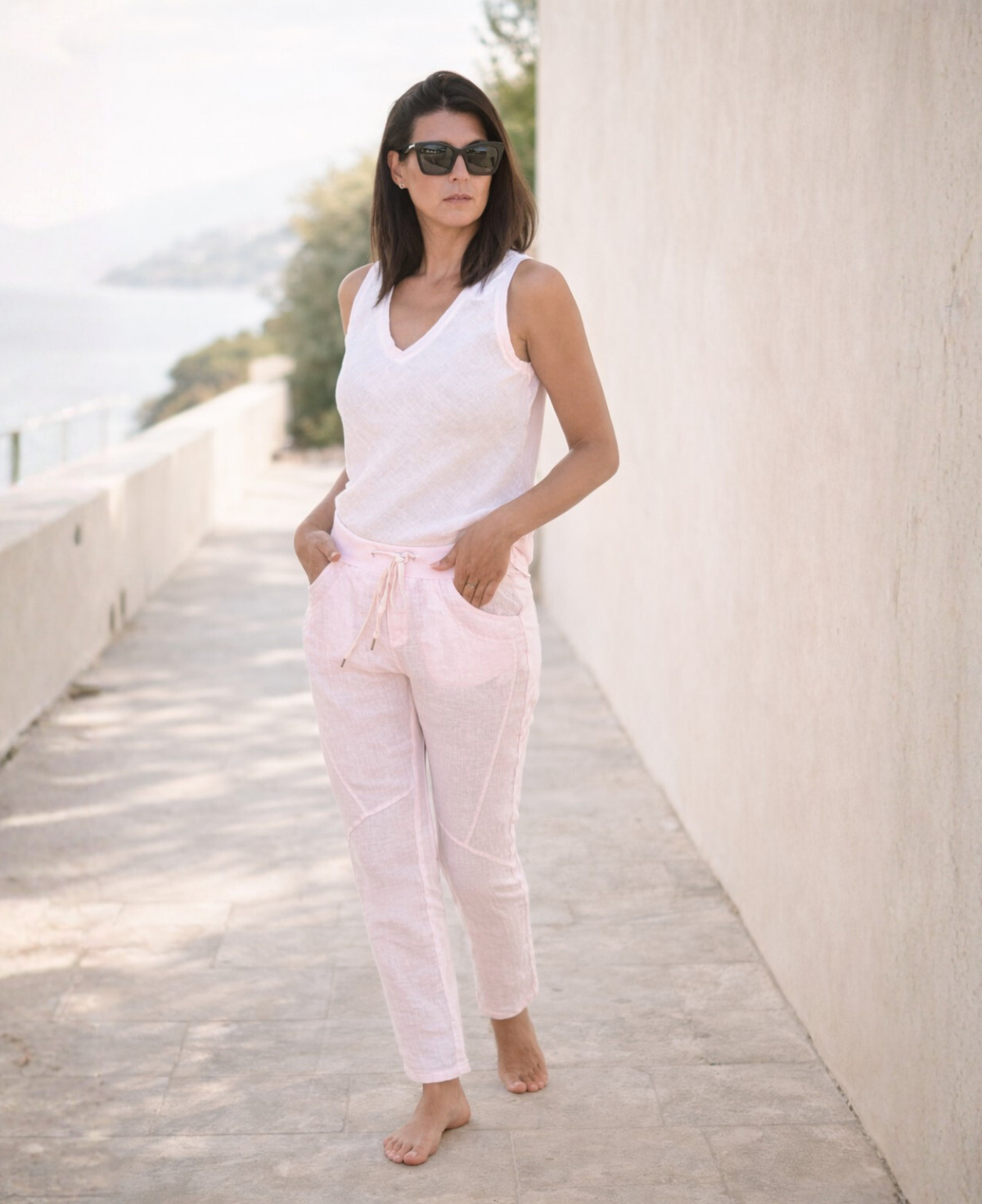 Full body editorial image of Jordan Linen Pants in blush worn on a tranquil coastal walk, showing natural linen movement.