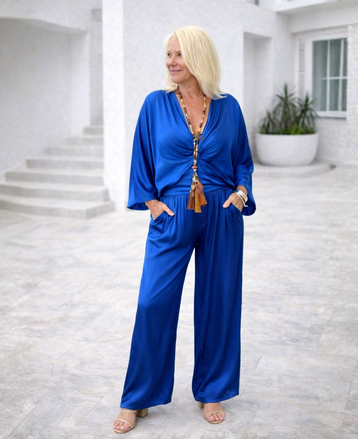 Woman wearing electric blue Juliette satin pants with fluid drape, shown from a front three-quarter angle on a Mediterranean terrace