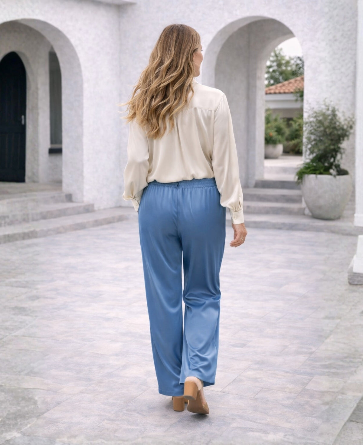 Back angled view of blue-teal Juliette satin pants showing satin sheen and flowing silhouette on a Mediterranean terrace