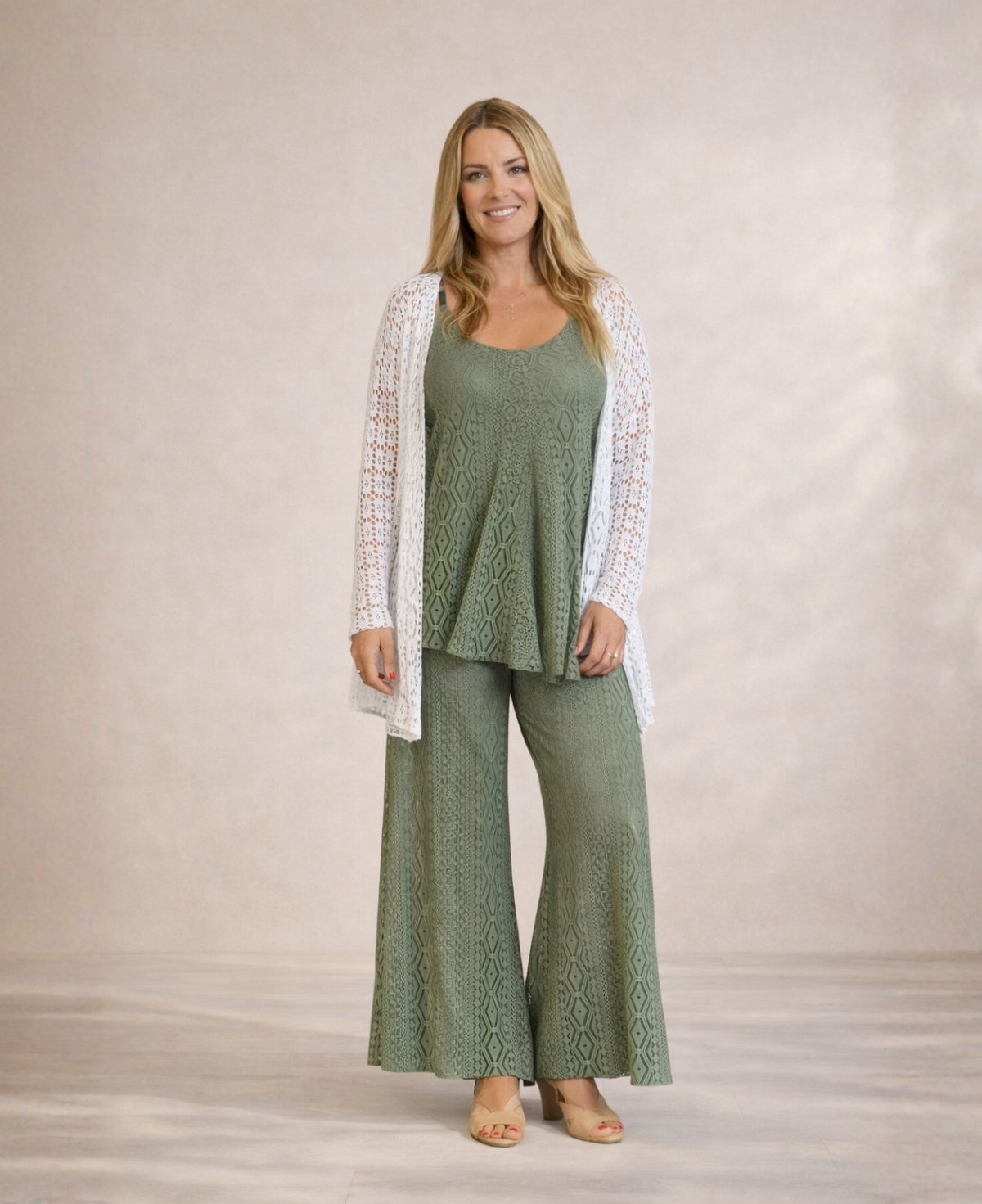 Full length studio shot of Louella Lace Cardi in White worn over green lace top and pants against warm sand backdrop.