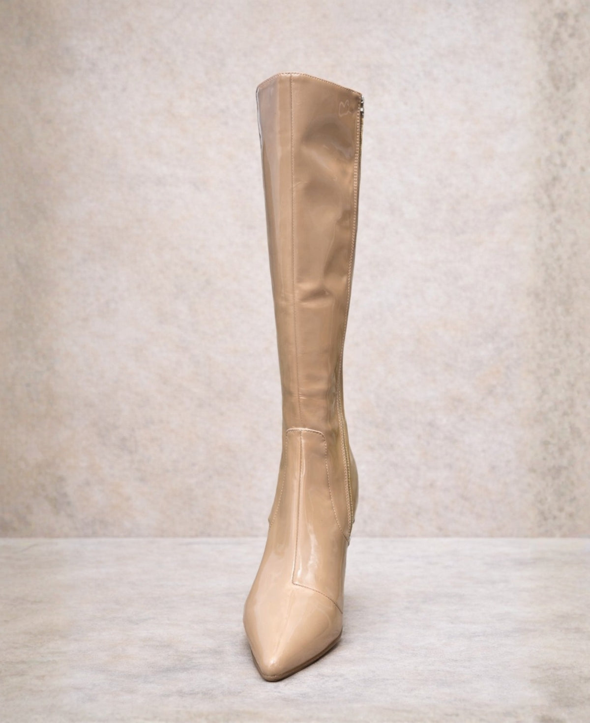 Front angled view of dark nude patent knee high boots with glossy finish and pointed toe on stone studio background