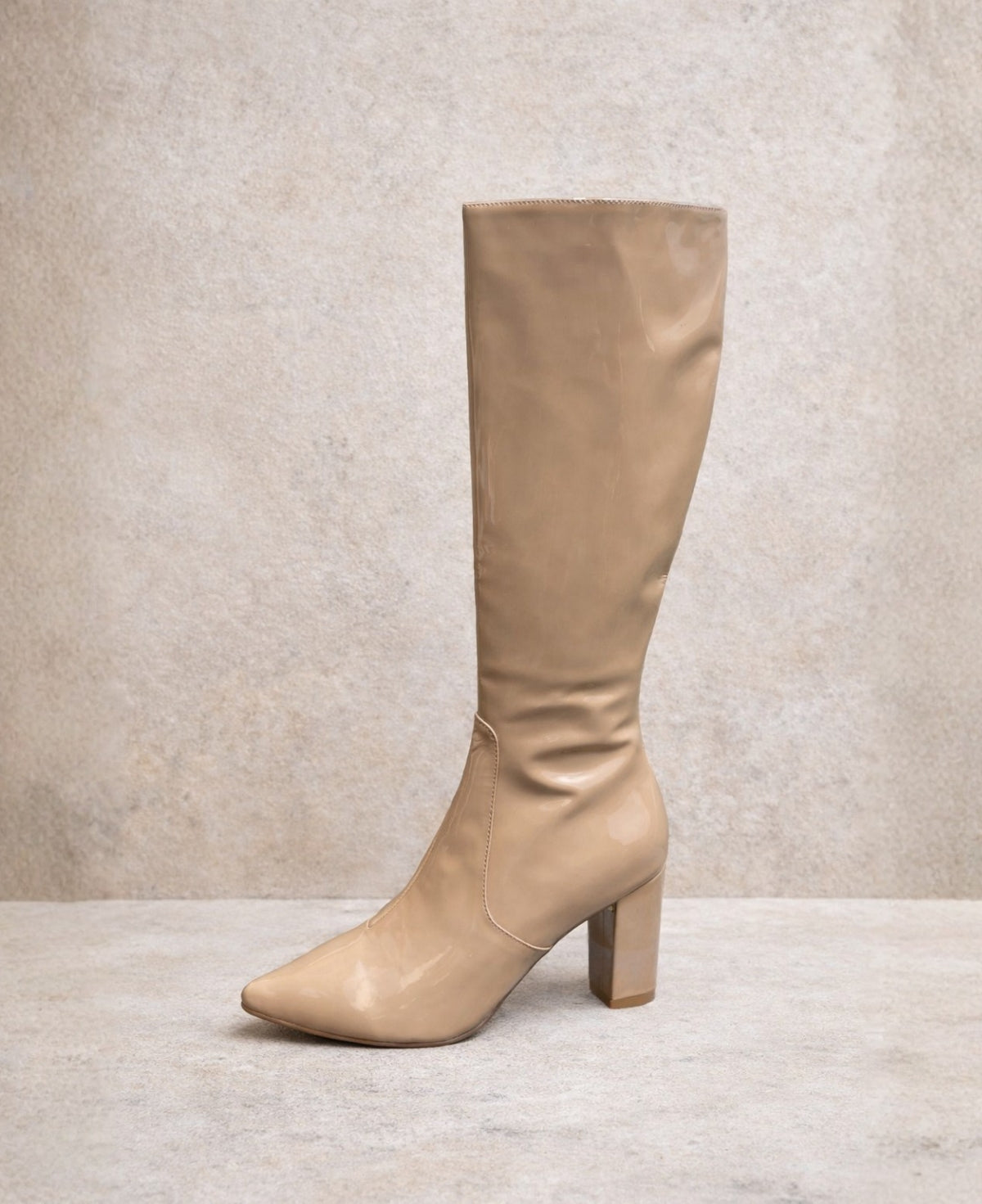 Dark nude patent knee high boots with pointed toe and block heel photographed against a soft stone studio background