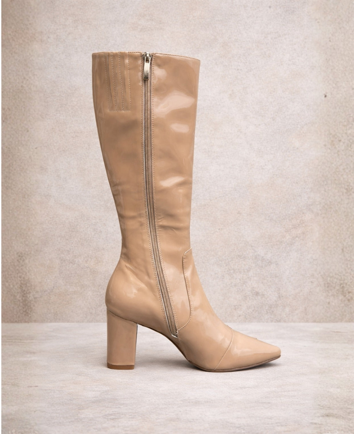 Dark nude patent knee high boots showing side zip fastening, pointed toe and mid block heel on neutral stone background