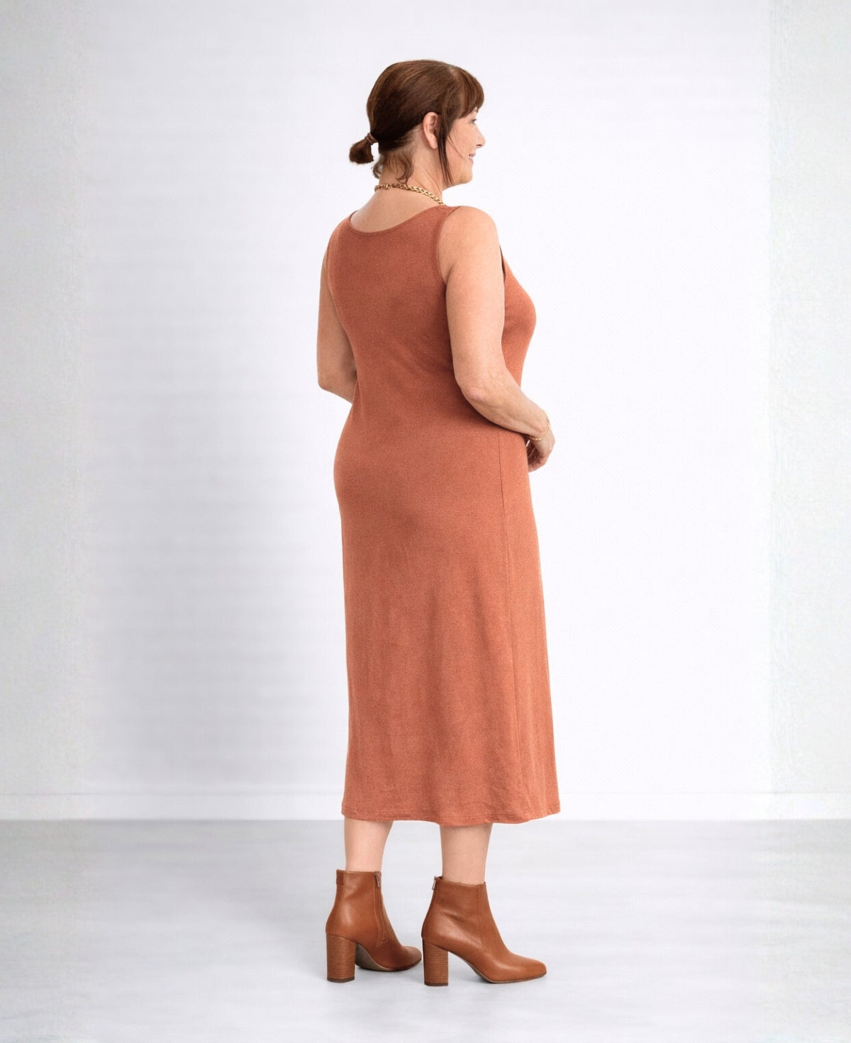 Back angled view of the Nell Knit Dress in copper tone styled with tan leather ankle boots on mature model in studio setting.
