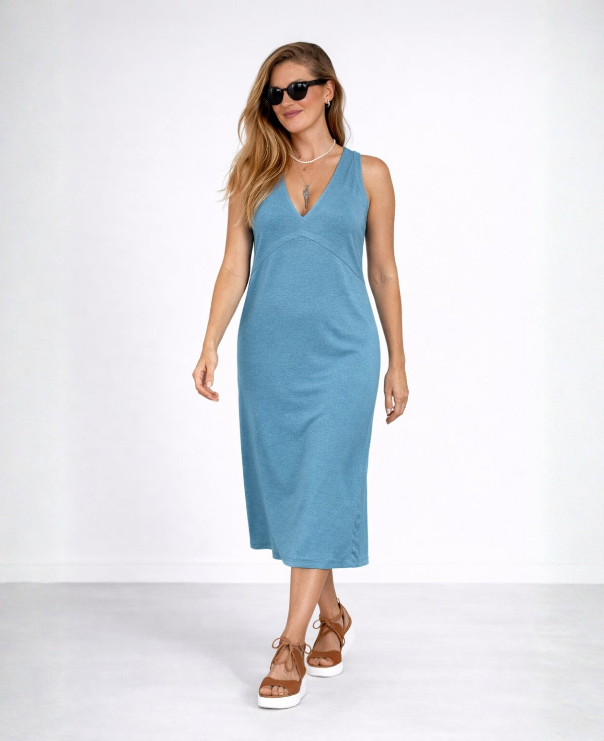 Model walking in the Nell Knit Dress in teal blue wearing sunglasses, showing natural drape and midi length in studio setting.
