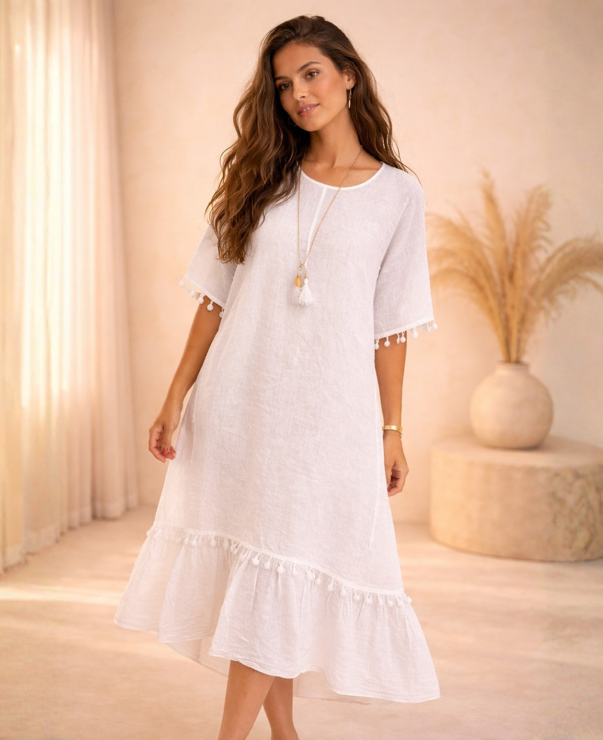Front angled view of the Paradiso Kenya Linen Dress in white linen, highlighting the relaxed cut, tassel sleeve detail, tiered hem, and natural drape in a warm studio setting.