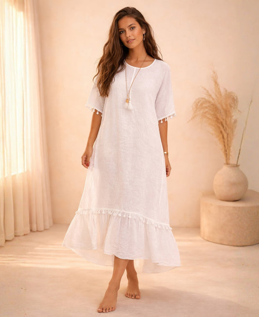 Model wearing the Paradiso Kenya Linen Dress in white, featuring a relaxed fit, short sleeves with tassel trim, and a tiered hem, photographed in a warm minimalist studio.