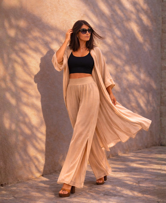 Editorial image of Pippa Silk Pants in Caramel silk blend, styled with black cropped tank top and flowing matching duster, photographed in warm golden hour light against textured wall.