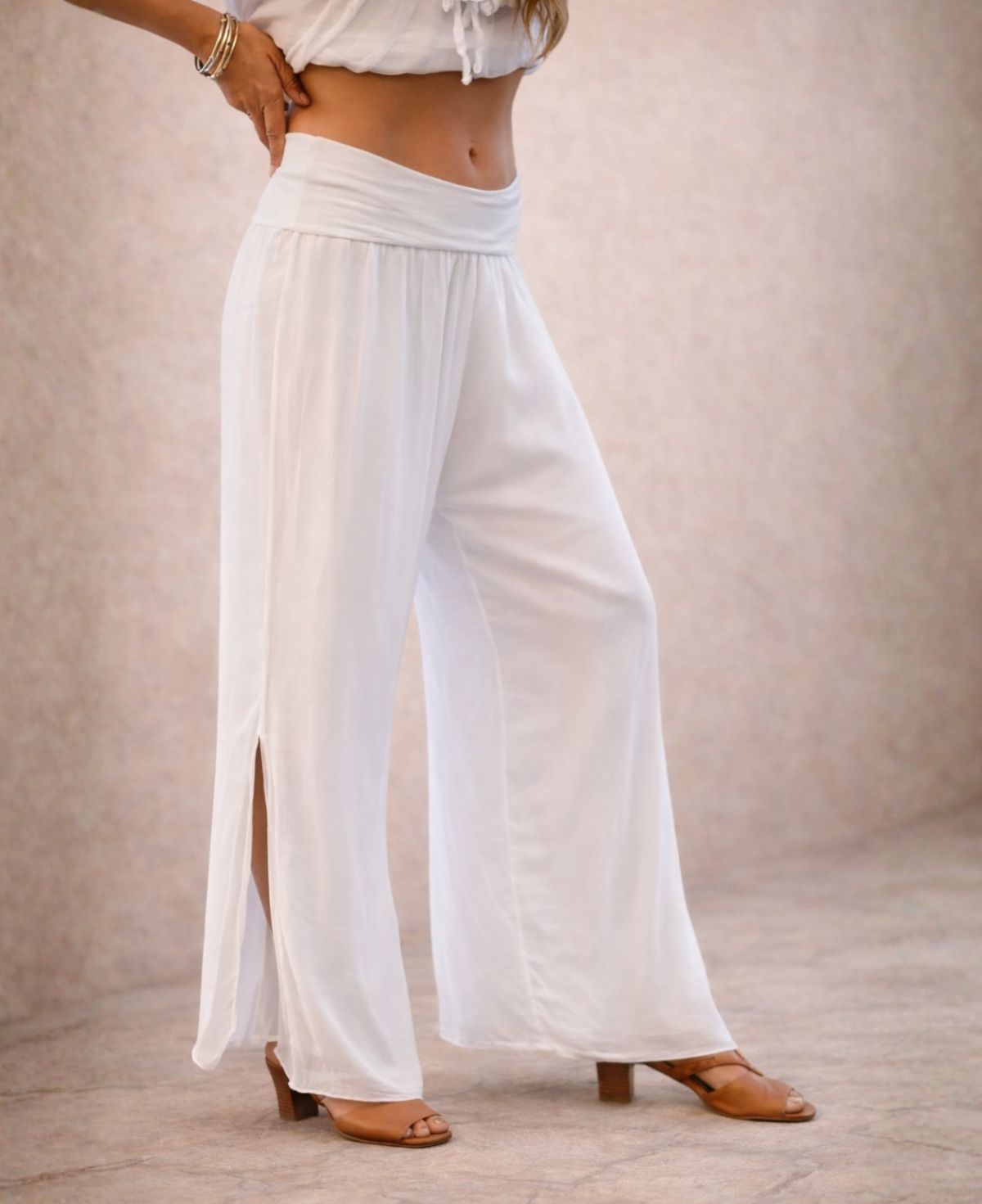 Close-up studio detail of Pippa Silk Pants in optic white silk blend, showing wide-leg shape, soft drape, elastic waistband and knee-high side slit in a warm stone background.