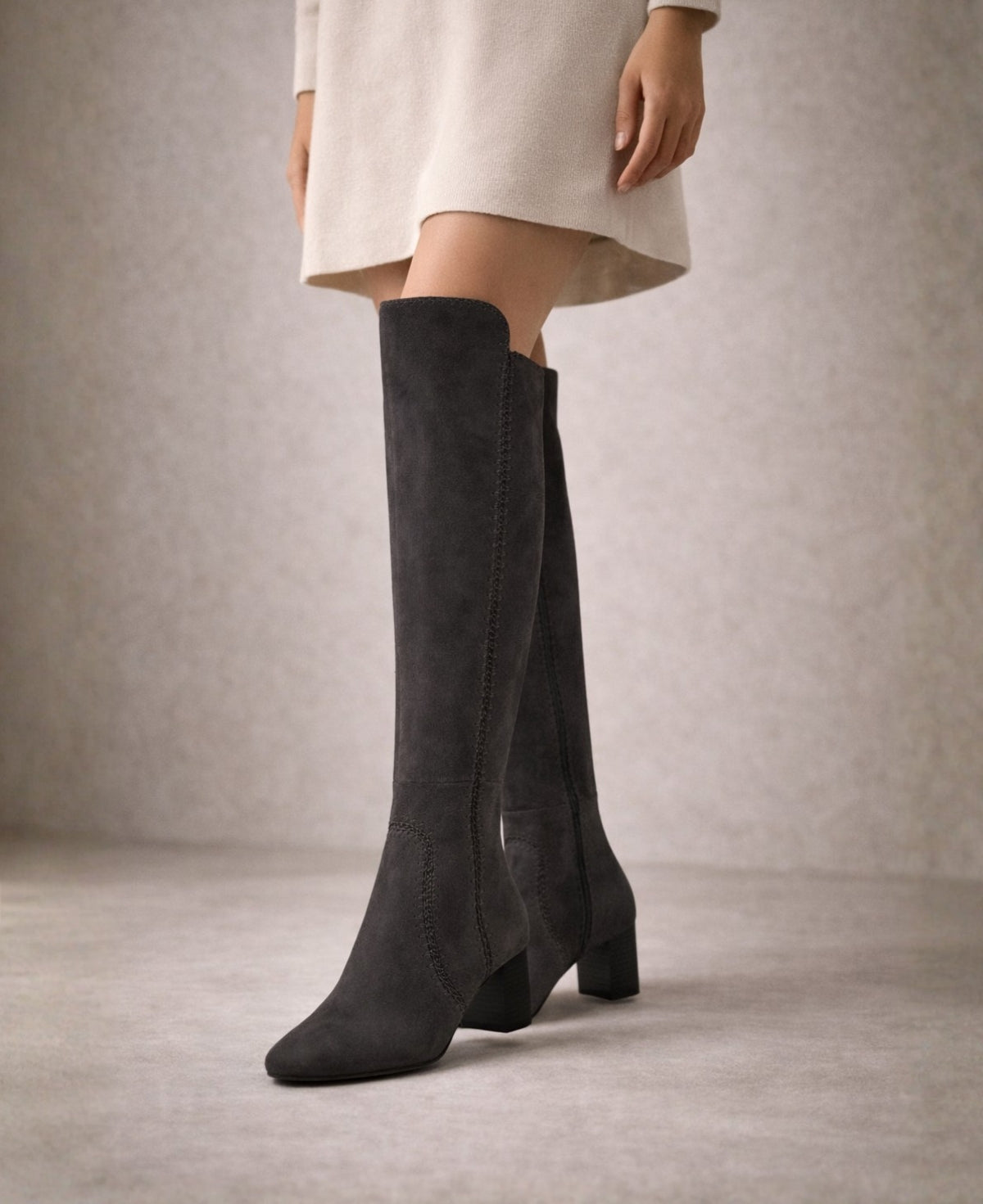 Model wearing black swept suede knee high boots shown from a side angle, highlighting the higher front rise and elegant block heel in a minimalist studio setting
