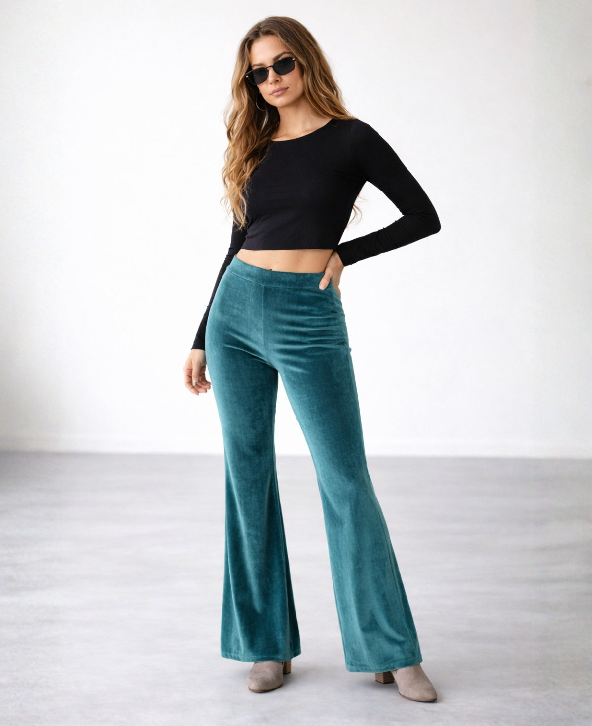 Model wearing teal velour flare pants with flared leg and flat waistband, styled with black top and boots, studio shot