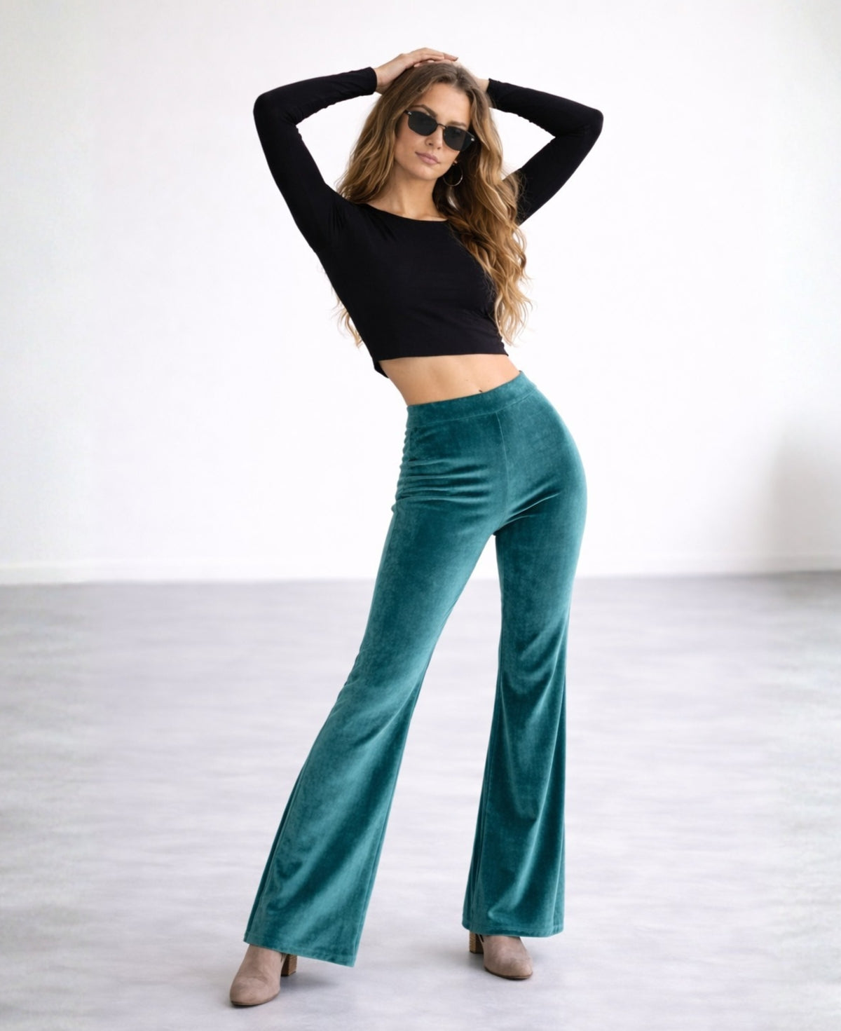 Model wearing teal velour flare pants with a flat waistband, styled with a black cropped top and ankle boots, full body studio shot showing the wide leg silhouette and soft sheen of the fabric