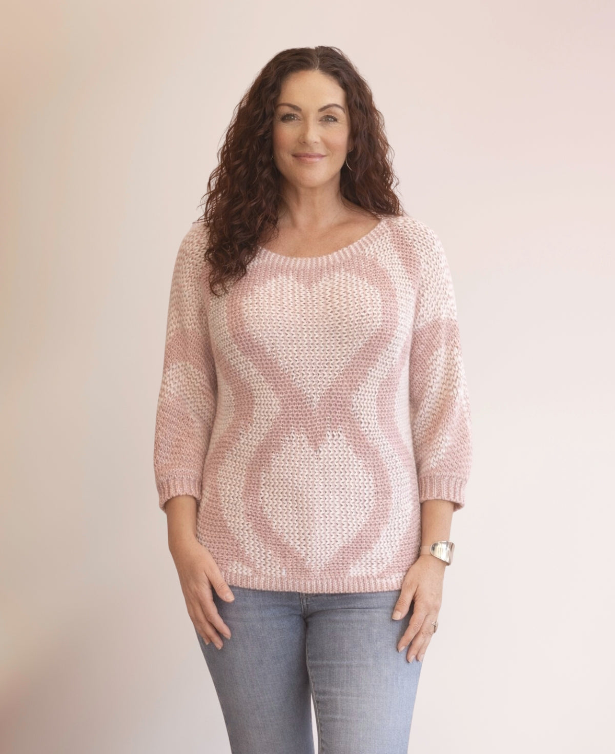 Model wearing the Valentine Wool Blend Jumper in Blush with soft pink heart knit pattern, styled with blue jeans against a warm oatmeal studio background.