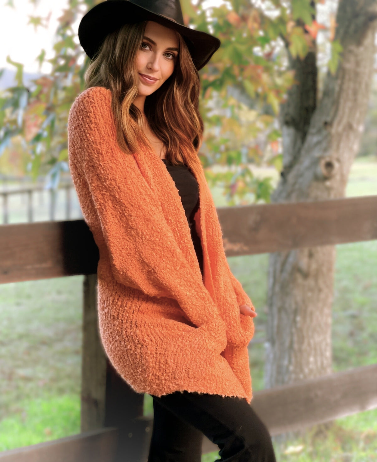 Woman wearing orange bouclé open-front cardigan styled with black outfit and wide-brim hat, photographed outdoors against rustic fence and autumn trees.