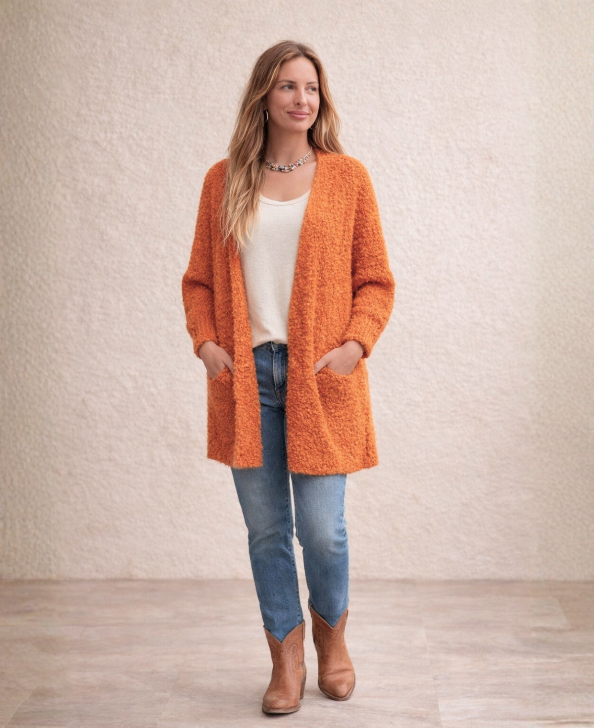 Woman wearing orange bouclé open-front cardigan with cream top, mid-wash jeans and tan leather cowboy boots against cream stucco wall.