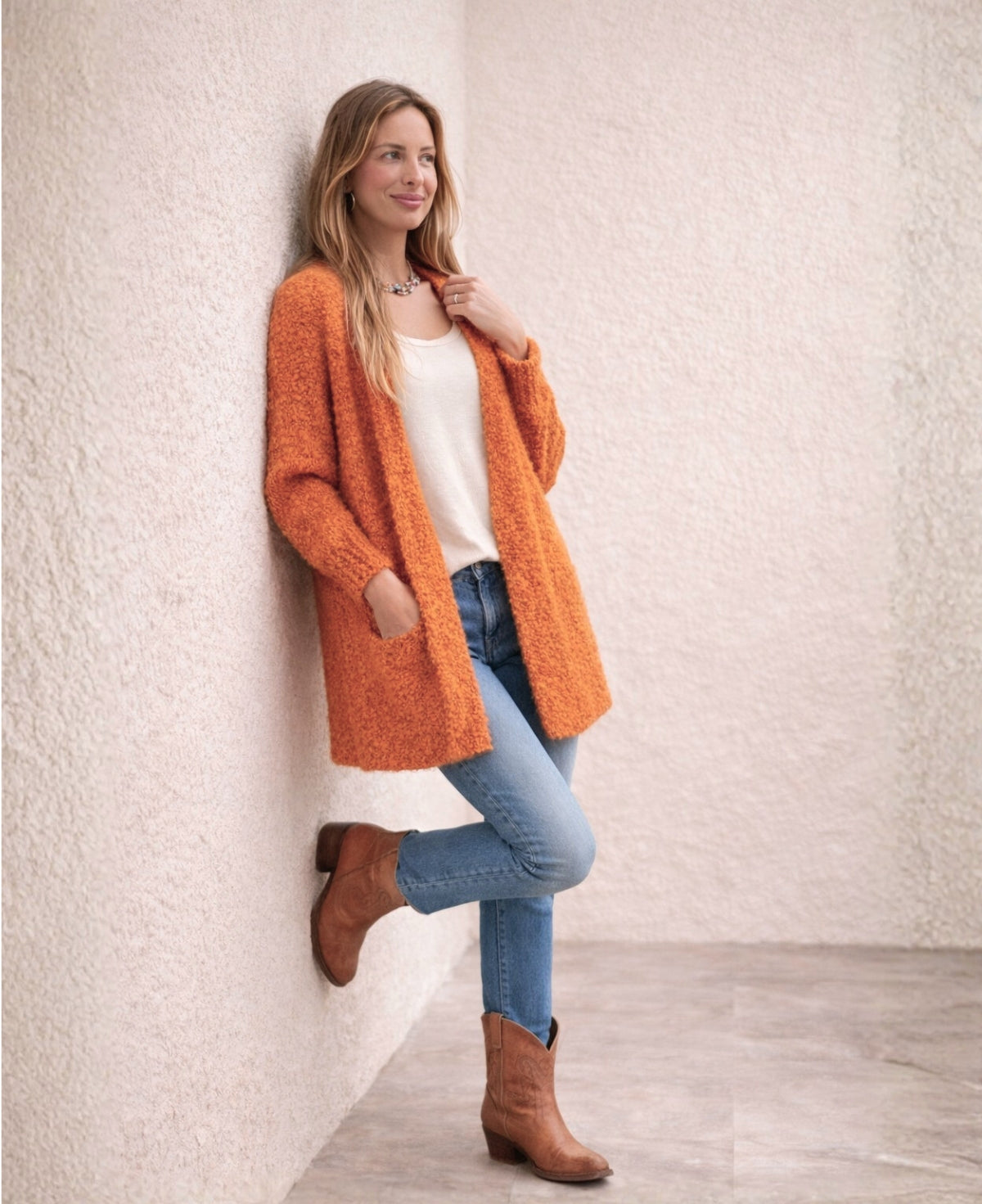 Side view of orange textured bouclé cardigan styled with cream base layer, straight denim and natural leather cowboy boots against neutral stucco wall.