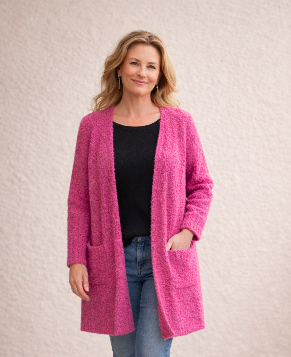 Woman wearing vibrant pink bouclé open-front cardigan with black top, mid-wash straight jeans and black ankle boots against cream stucco wall.