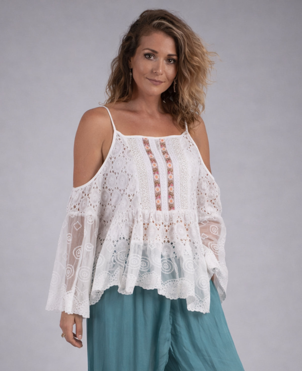 Woman wearing a white lightweight lace summer flowy top with cold shoulder sleeves and embroidered front panel, styled with teal loose pants against a soft grey studio background