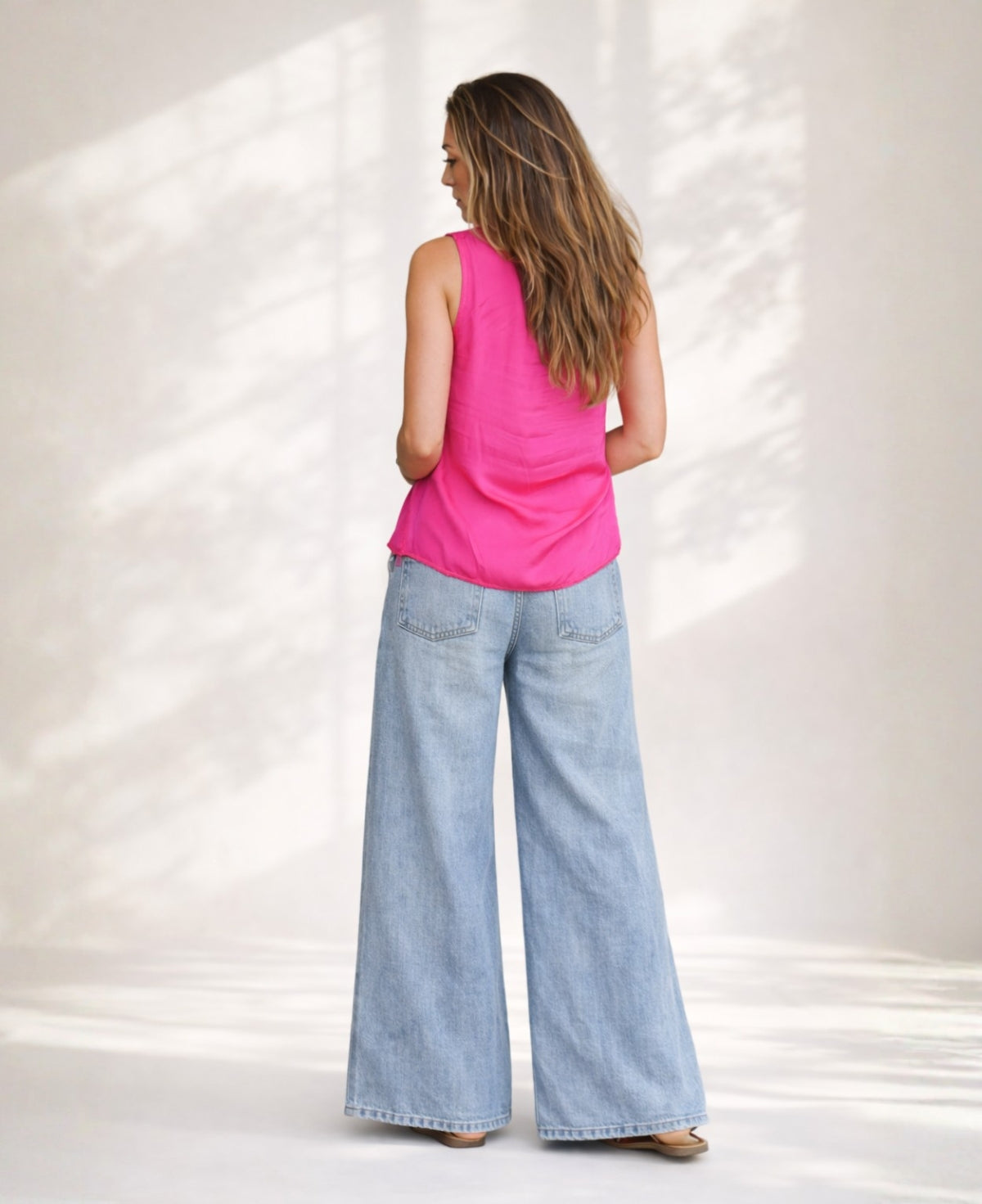 Back view of hot pink satin sleeveless top with ruffle neckline styled with wide leg denim jeans in soft studio background