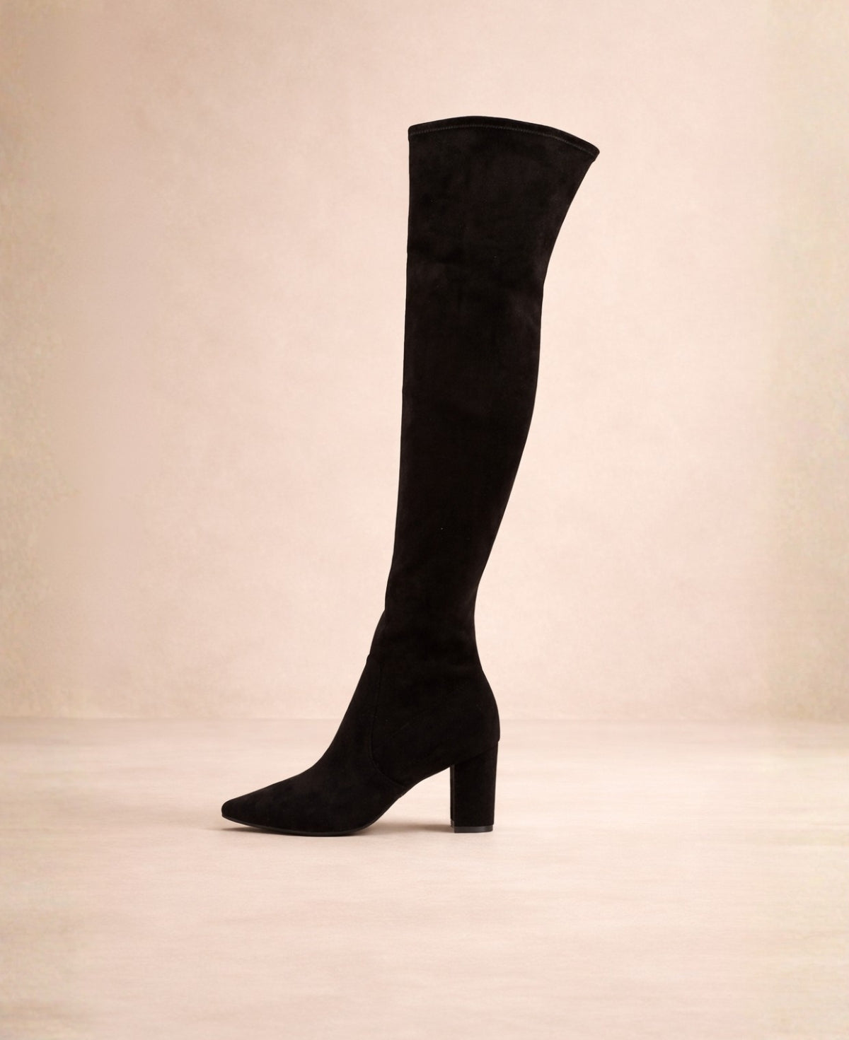 Black microsuede above-the-knee boots with pointed toe and block heel shown from the outer side in a warm studio background.