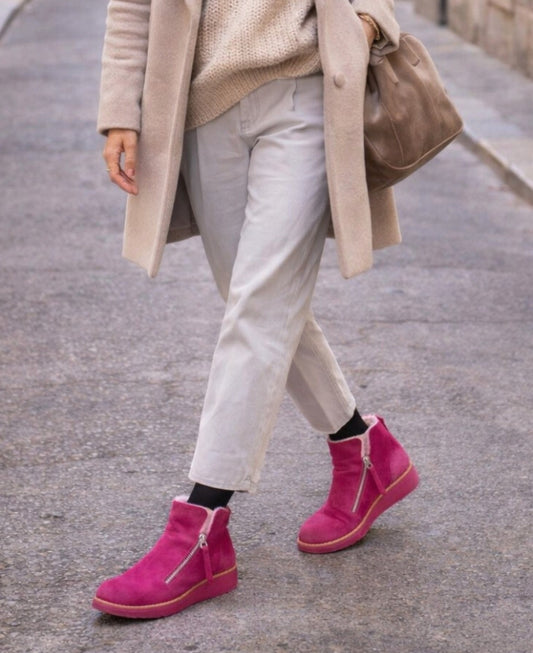 Woman wearing Opal fuchsia suede ankle boots styled with neutral tones while walking through a quiet city street.